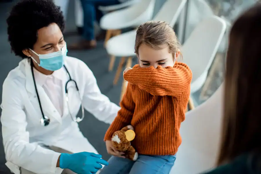 A masked healthcare provider speaking to a little girl in an urgent care who’s coughing into her elbow.