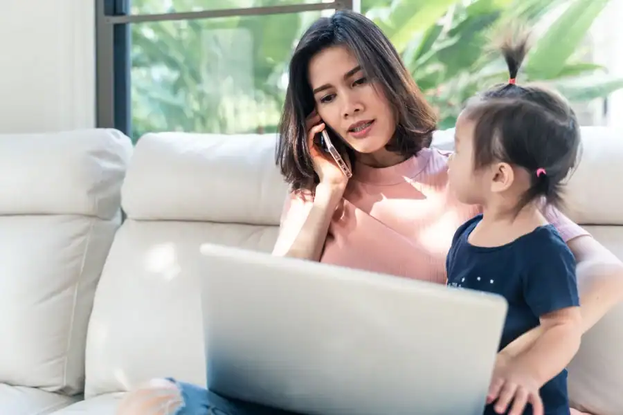 A mother looking at a laptop and talking on the phone with her young daughter on her lap.