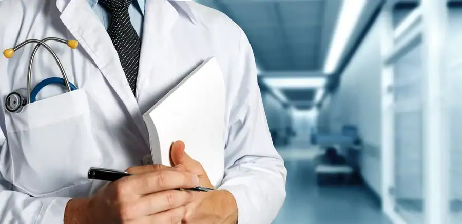 A doctor wearing a white coat with a stethoscope in the pocket and holding a notepad and pen.