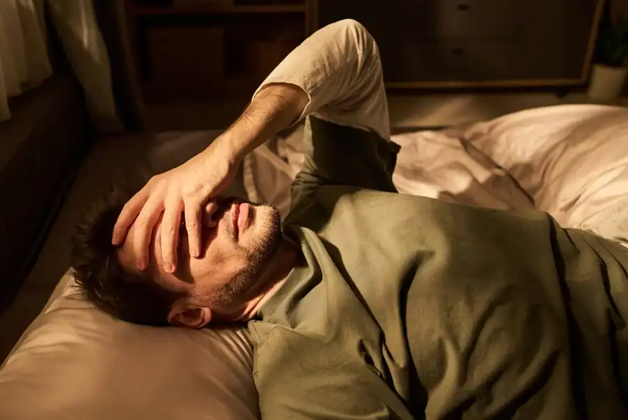A man with a morning headache lying in bed with his hand over his eyes.